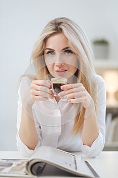 A young beautiful Caucasian blonde woman rests at home. A woman sits at a table, drinking coffee from a glass cup and