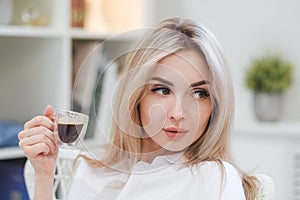 A young beautiful Caucasian blonde woman rests at home. A woman sits at a table, drinking coffee from a glass cup and