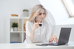 Young beautiful caucasian blonde woman working on laptop at home and drinking coffee. Remote work or freelance.