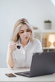 Young beautiful caucasian blonde woman working on laptop at home and drinking coffee. Remote work or freelance.