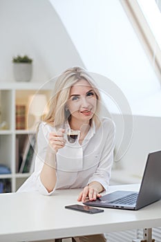 Young beautiful caucasian blonde woman working on laptop at home and drinking coffee. Remote work or freelance.
