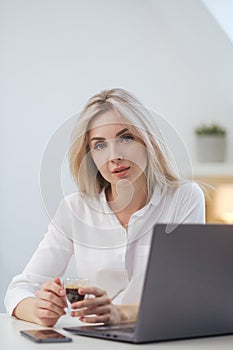 Young beautiful caucasian blonde woman working on laptop at home and drinking coffee. Remote work or freelance.