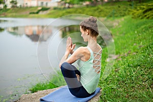 Young beautiful caucasian brunette girl doing yoga on a green lawn against the background of the river