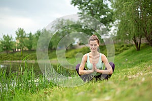 Young beautiful caucasian brunette girl doing yoga on a green lawn against the background of the river