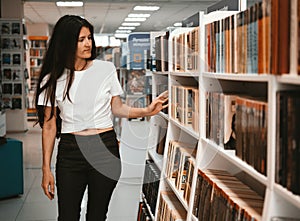 Young beautiful caucasian brunette woman in  white T-shirt chooses books between the  bookshelves