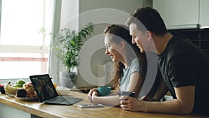 Young beautiful caucasian couple sitting in modern kitchen at table in front of laptop skyping with two girls, they are