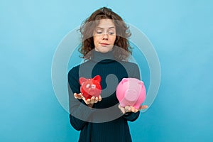 Young and beautiful caucasian girl with curly hair keeps small red pig moneybox, big pink pig moneybox and smiles