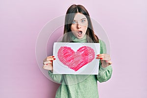 Young beautiful caucasian girl holding heart draw afraid and shocked with surprise and amazed expression, fear and excited face