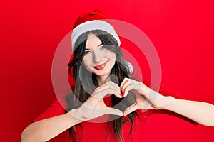 Young beautiful caucasian girl wearing christmas hat smiling in love showing heart symbol and shape with hands