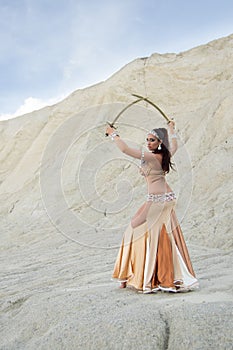 Young beautiful Caucasian woman belly dancer posing in desert with swords, back side