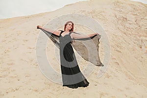 Young beautiful Caucasian woman in black chiffon dress posing in desert landscape with sand.