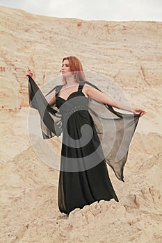 Young beautiful Caucasian woman in black chiffon dress posing in desert landscape with sand.