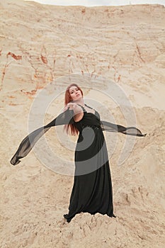 Young beautiful Caucasian woman in black chiffon dress posing in desert landscape with sand.