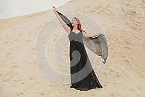 Young beautiful Caucasian woman in black chiffon dress posing in desert landscape with sand.