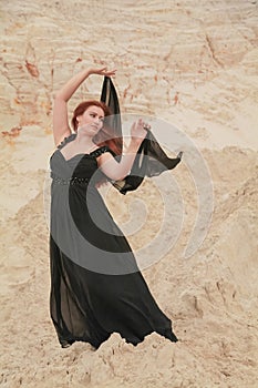 Young beautiful Caucasian woman in black chiffon dress posing in desert landscape with sand.