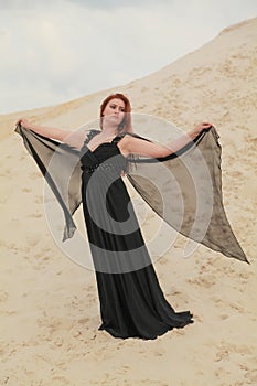 Young beautiful Caucasian woman in black chiffon dress posing in desert landscape with sand.