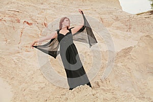 Young beautiful Caucasian woman in black chiffon dress posing in desert landscape with sand.