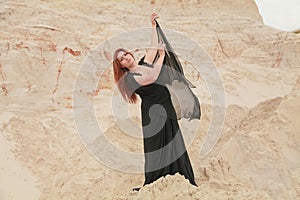 Young beautiful Caucasian woman in black chiffon dress posing in desert landscape with sand.