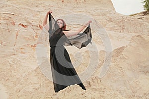 Young beautiful Caucasian woman in black chiffon dress posing in desert landscape with sand.
