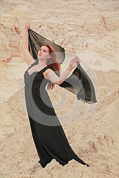 Young beautiful Caucasian woman in black chiffon dress posing in desert landscape with sand.