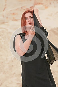 Young beautiful Caucasian woman in black chiffon dress posing in desert landscape with sand.