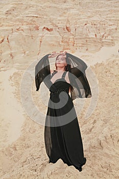 Young beautiful Caucasian woman in black chiffon dress posing in desert landscape with sand.