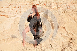 Young beautiful Caucasian woman in black sexy long dress posing in desert landscape with sand.