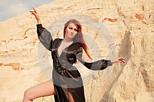 Young beautiful Caucasian woman in black sexy long dress posing in desert landscape with sand.