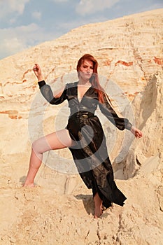 Young beautiful Caucasian woman in black sexy long dress posing in desert landscape with sand.