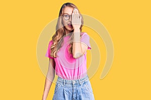 Young beautiful caucasian woman with blond hair wearing casual clothes and glasses covering one eye with hand, confident smile on