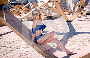 Blonde Caucasian Woman on the Beach With Blue Flounce Top  Swimsuit, Leaning on a Wood Structure and Looking Away photo