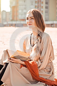 Young beautiful caucasian woman in boho dress and red thin scarf sits on beach in city and reads book and dreaming