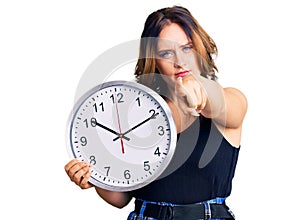 Young beautiful caucasian woman holding big clock pointing with finger to the camera and to you, confident gesture looking serious