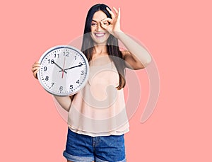 Young beautiful caucasian woman holding big clock smiling happy doing ok sign with hand on eye looking through fingers