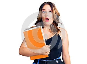 Young beautiful caucasian woman holding book scared and amazed with open mouth for surprise, disbelief face