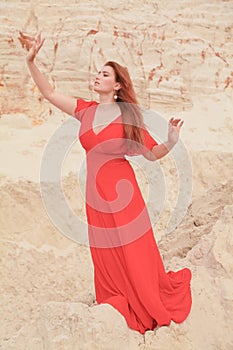 Young beautiful Caucasian woman in long red dress posing in desert landscape with sand.