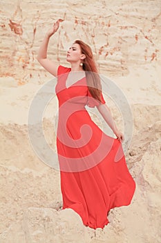 Young beautiful Caucasian woman in long red dress posing in desert landscape with sand.