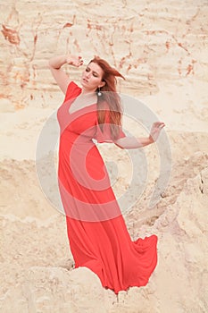 Young beautiful Caucasian woman in long red dress posing in desert landscape with sand.