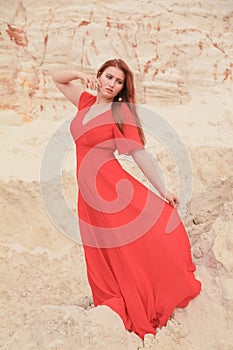 Young beautiful Caucasian woman in long red dress posing in desert landscape with sand.