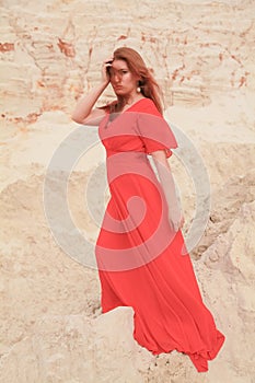 Young beautiful Caucasian woman in long red dress posing in desert landscape with sand.