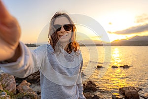 Young beautiful caucasian woman make a self portrait holding the camera at sunset on sea rocks - Smartphone selfie with water