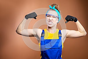Young beautiful caucasian woman repairman worker with blue eyes in uniform showing arm muscles, proudly smiling and