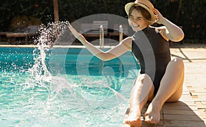 young beautiful caucasian woman sits in the summer on the shore of the pool in a straw hat and makes splashes of water