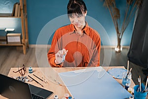 a young beautiful caucasian woman sits at a table at home and paints a picture on canvas with acrylic paints, watches an