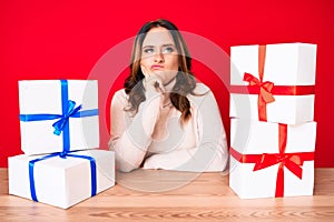 Young beautiful caucasian woman sitting on the table with presents thinking concentrated about doubt with finger on chin and