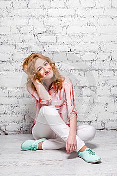 Young beautiful caucasian woman student is sitting on the floor near a white brick wall. Girl with red long curly hair