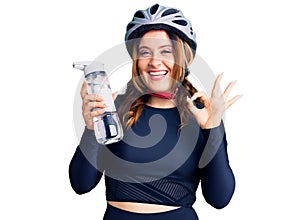 Young beautiful caucasian woman wearing bike helmet and holding water bottle doing ok sign with fingers, smiling friendly