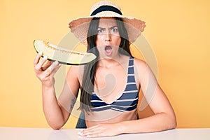 Young beautiful caucasian woman wearing bikini and hat holding slice of melon scared and amazed with open mouth for surprise,