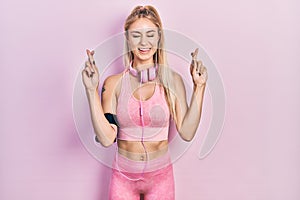 Young beautiful caucasian woman wearing gym clothes and using headphones gesturing finger crossed smiling with hope and eyes