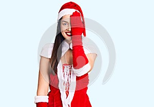 Young beautiful caucasian woman wearing santa claus costume covering one eye with hand, confident smile on face and surprise